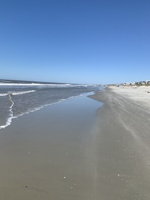 Een breed zandstrand met zachte golven, een helderblauwe lucht en een verre kustlijn met gebouwen.