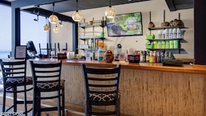 A bar area with wooden bar counter, two black chairs with patterned cushions, hanging spherical light fixtures, shelves with bottles and glasses, and a television mounted on the wall. - Beachfront, 24th Floor (Panama City Beach)