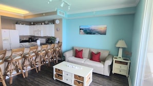 A living room with a sofa, coffee table, and a dining area with chairs and a kitchen in the background. - Beachfront, 24th Floor (Panama City Beach)