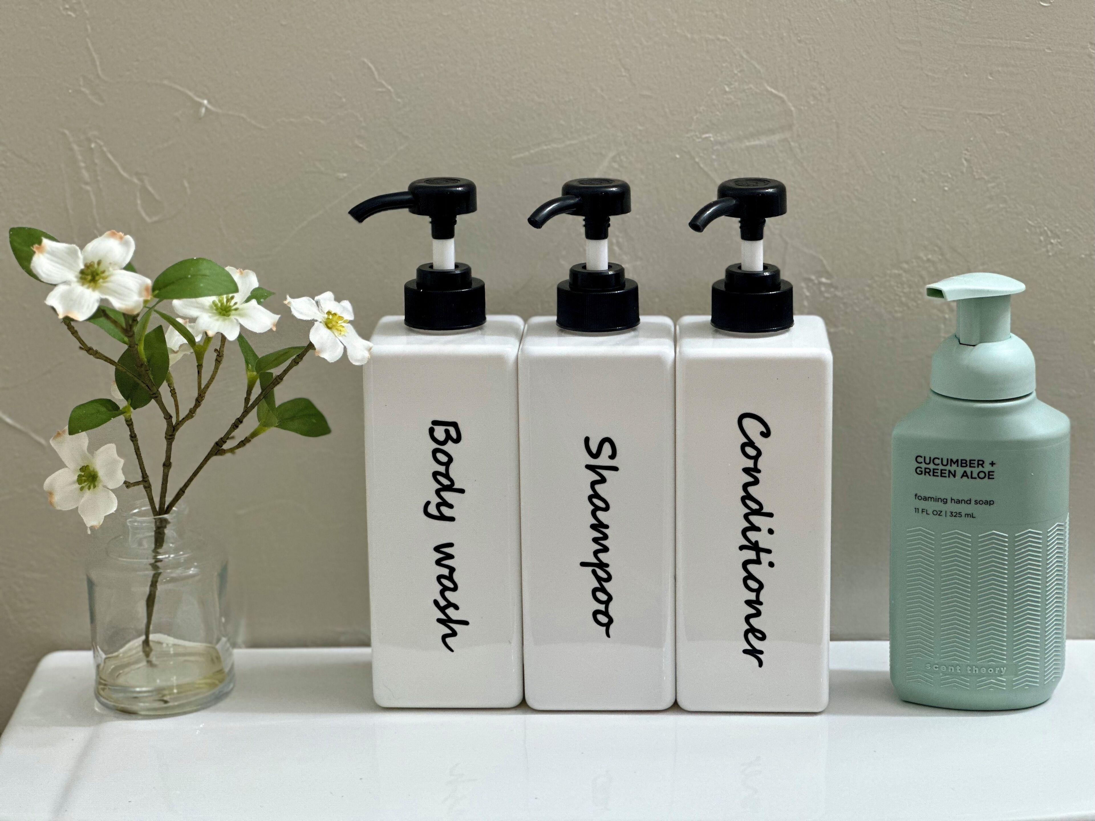 Trois flacons blancs étiquetés « Gel douche », « Shampoing » et « Après-shampoing », dotés de pompes noires, et un flacon vert étiqueté « Savon liquide pour les mains au concombre et à l’aloe vera », le tout posé sur une surface blanche.