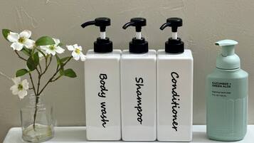Trois flacons blancs étiquetés « Gel douche », « Shampoing » et « Après-shampoing », dotés de pompes noires, et un flacon vert étiqueté « Savon liquide pour les mains au concombre et à l’aloe vera », le tout posé sur une surface blanche.