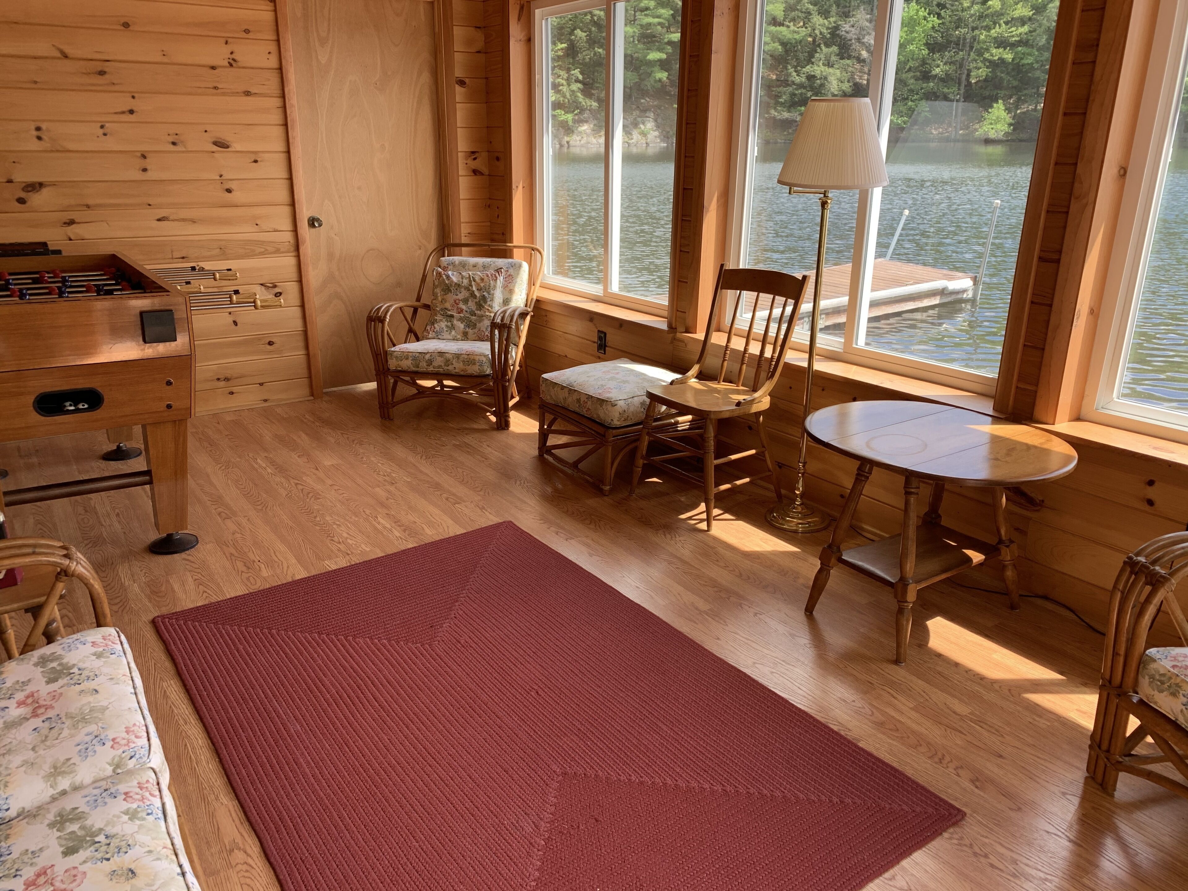 Interior de una cabaña de madera con un escritorio, sillas, una mesita y vistas a un lago a través de amplias ventanas.