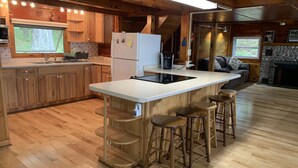 A modern kitchen with wooden cabinets, a central island with stools, a refrigerator, and a stone fireplace. - Lakefront Cottage near Adirondacks, Pontoon Boat, Sunroom, 2 Kayaks & Canoe (Hermon)