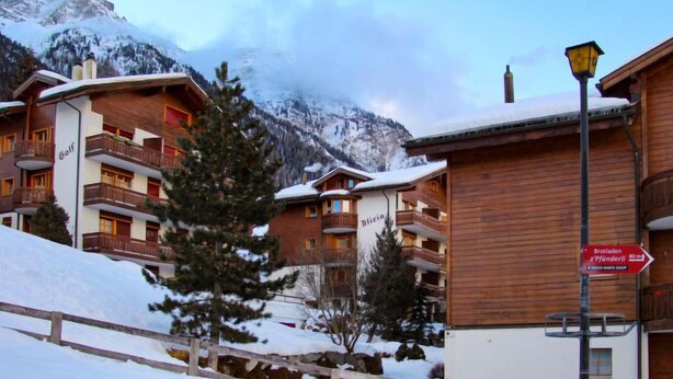 A snow-covered mountain resort with wooden buildings, balconies, and a sign indicating a distance to a shop. - Apartment Alicia by Interhome (Grächen)