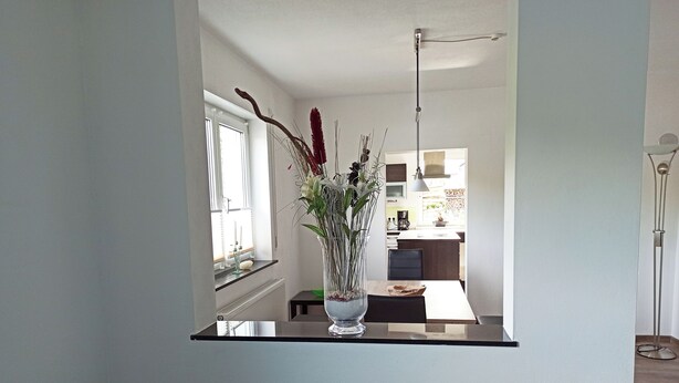 A modern kitchen with white cabinets, a dining area with a black chair, and a vase with flowers on a black table. - Urlaub in Ländlicher Umgebung (Illschwang)