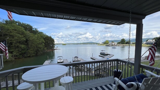 A covered deck with a table and chairs, overlooking a lake with boats and a dock.