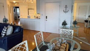 A modern kitchen with white cabinets, a dining area with a glass table and white chairs, and a living room with a dark sofa and decorative pillows.
