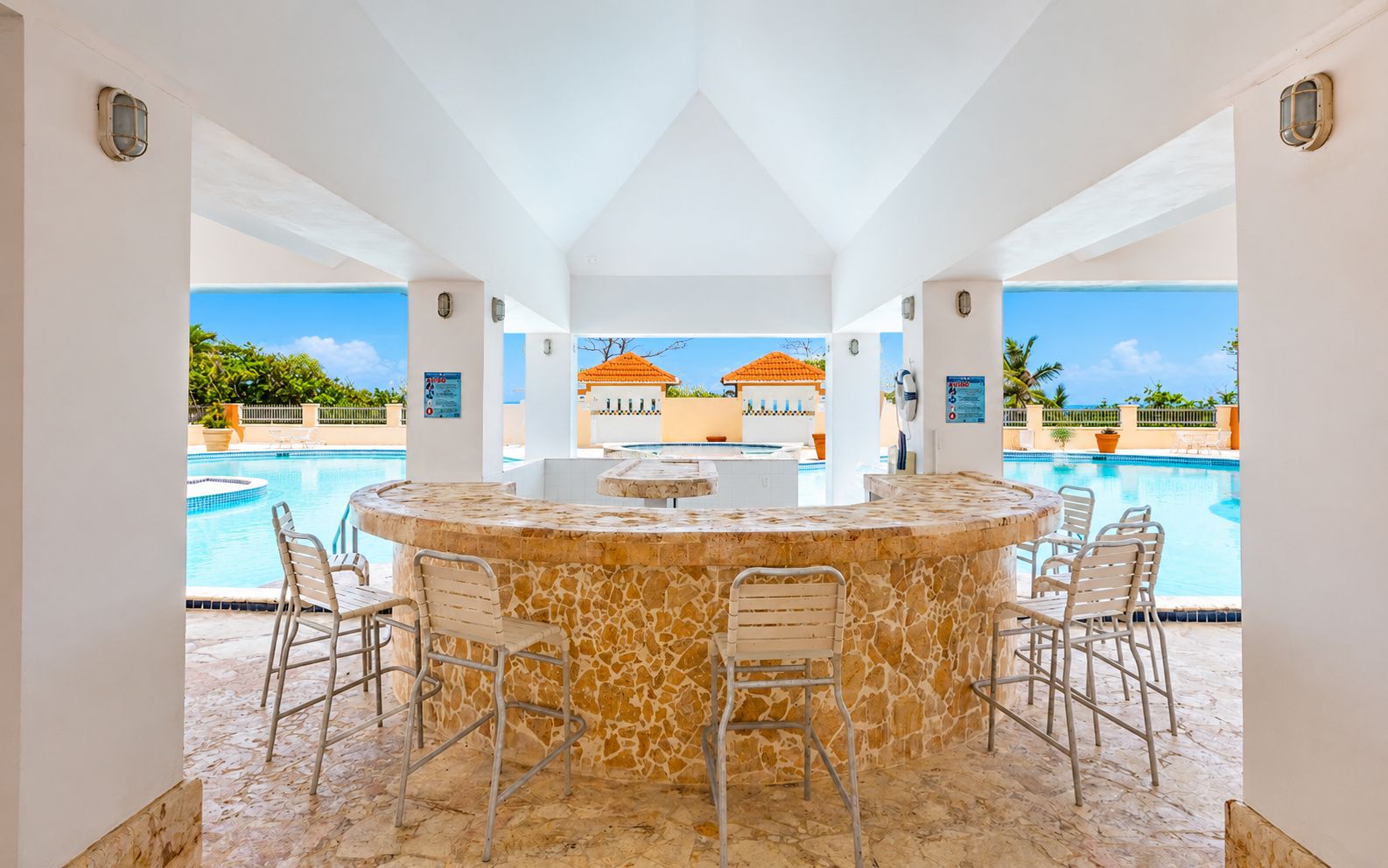 Un bar junto a la piscina con encimera de piedra y cuatro sillas, con vistas a una piscina y un edificio con tejado naranja.