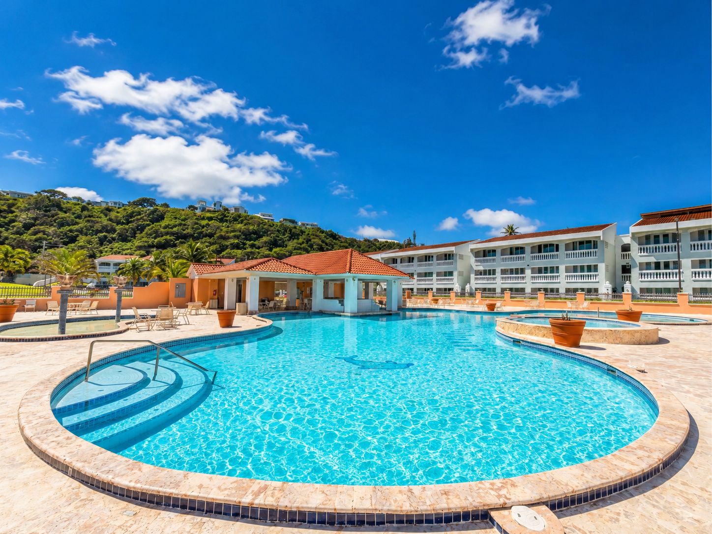 A hotel with a large swimming pool, a covered area, and a view of a hill with buildings.