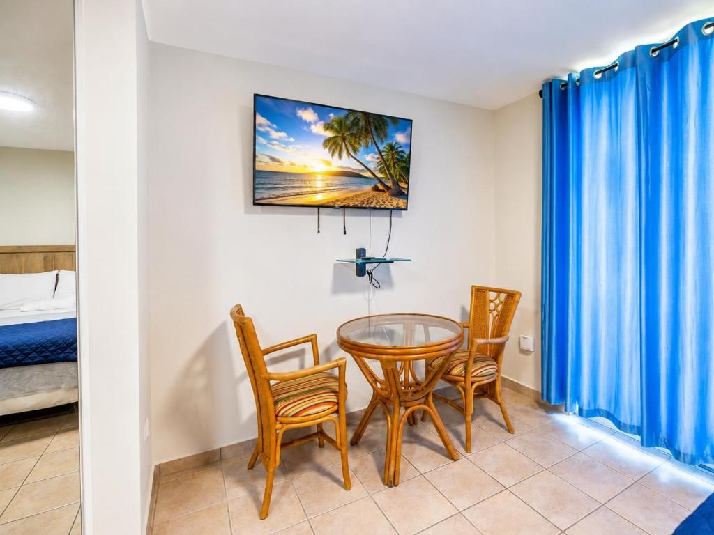 A room with a bed, a TV mounted on the wall, a dining table with chairs, and a window with blue curtains.