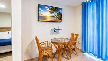 A room with a bed, a TV mounted on the wall, a dining table with chairs, and a window with blue curtains.