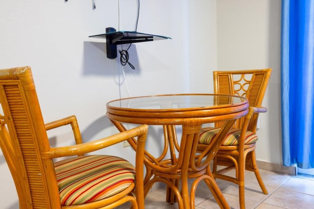Un comedor con sillas de mimbre y una mesa de vidrio, un enchufe en la pared y una cortina azul.