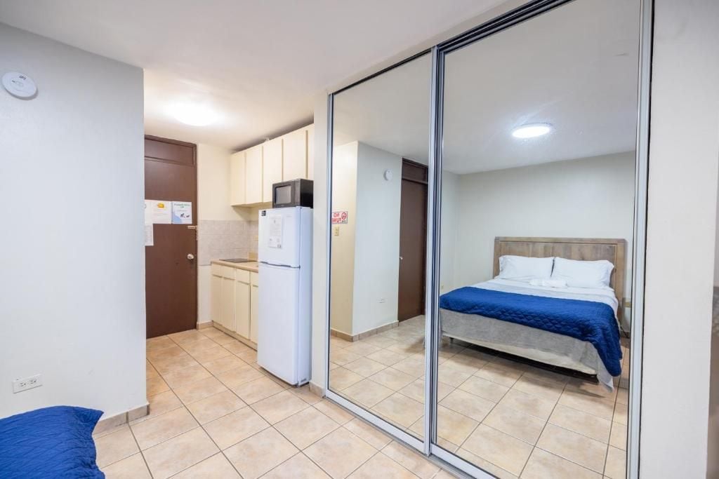 A compact room with a bed, a kitchenette, and a sliding door leading to another room.