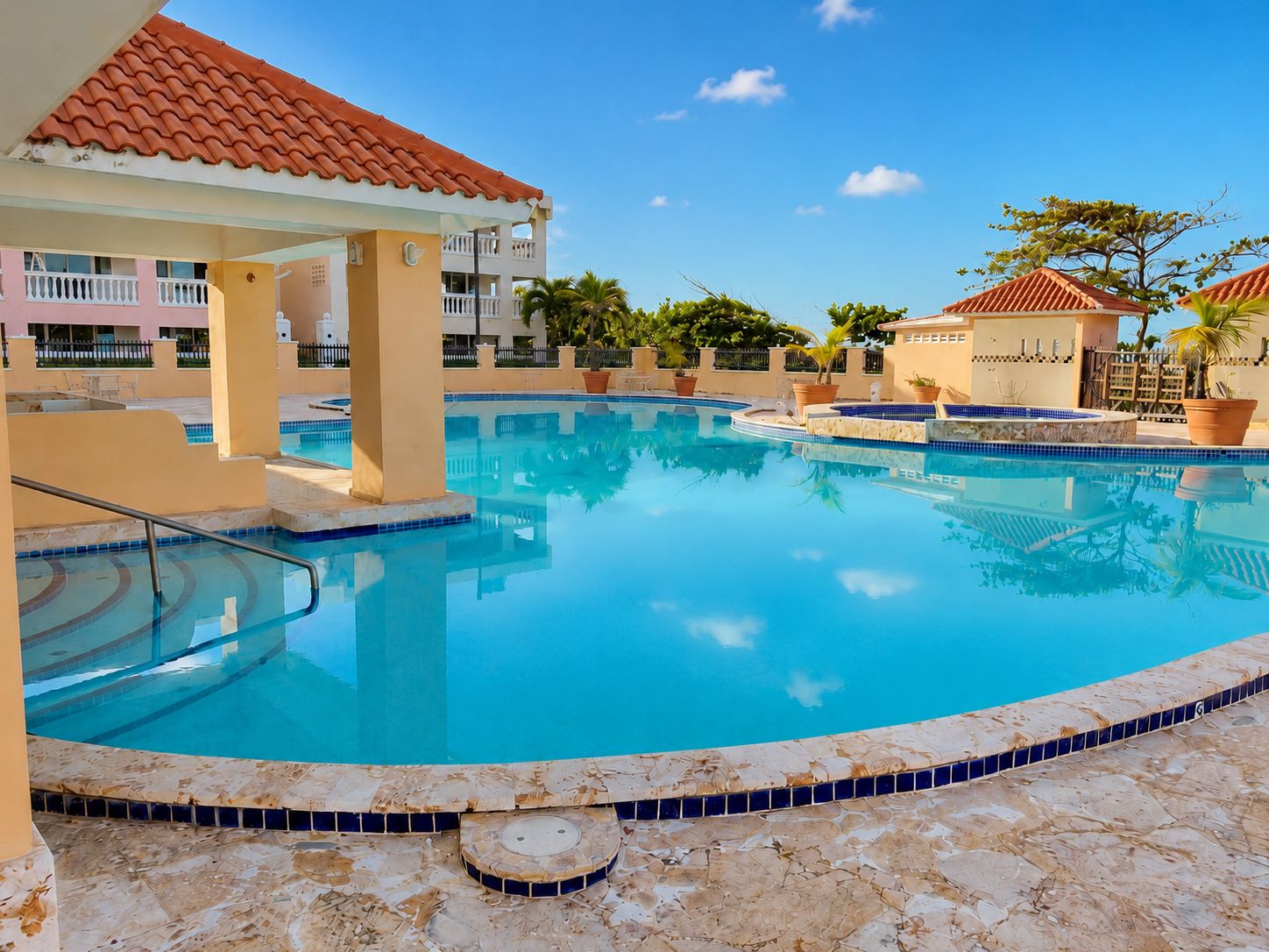 A swimming pool with a tiled edge, a covered seating area, and a building with a red-tiled roof in the background.