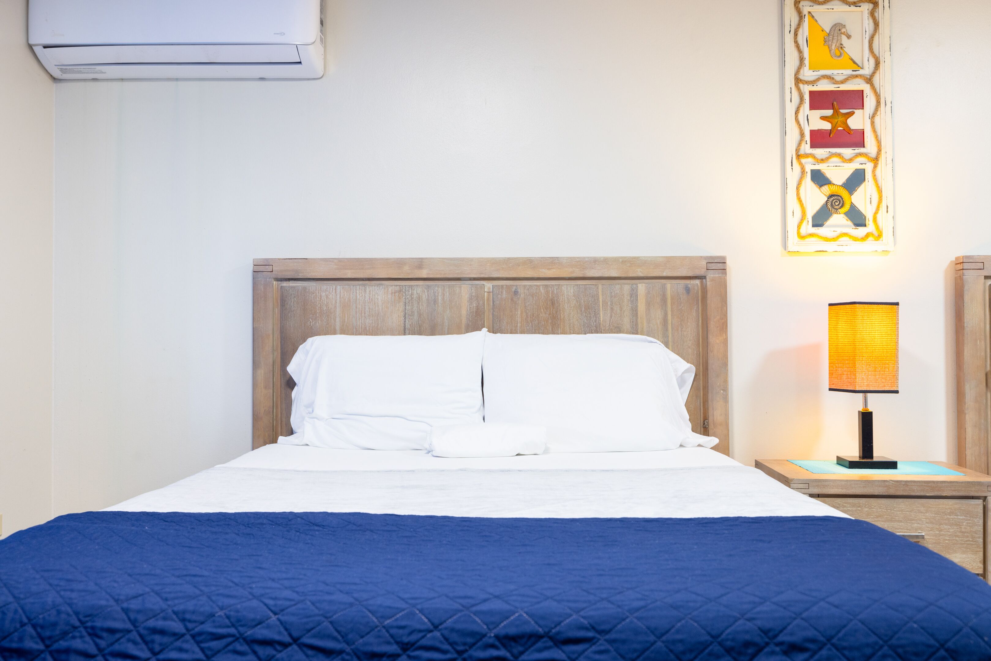 Een net opgemaakt bed met een blauwe quilt, een houten hoofdbord, een wandairconditioner en een decoratief wandobject.