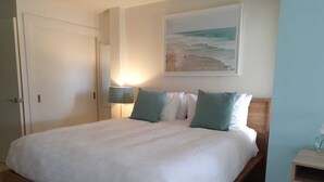 En pænt redt seng med et hvidt dynebetræk og to puder, en senget lampe og et billede i ramme af en strandscene på væggen.