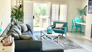A modern living room with a grey sectional sofa, a coffee table, and a teal armchair. There is a dining area with a table and chairs, and a large window with blinds.