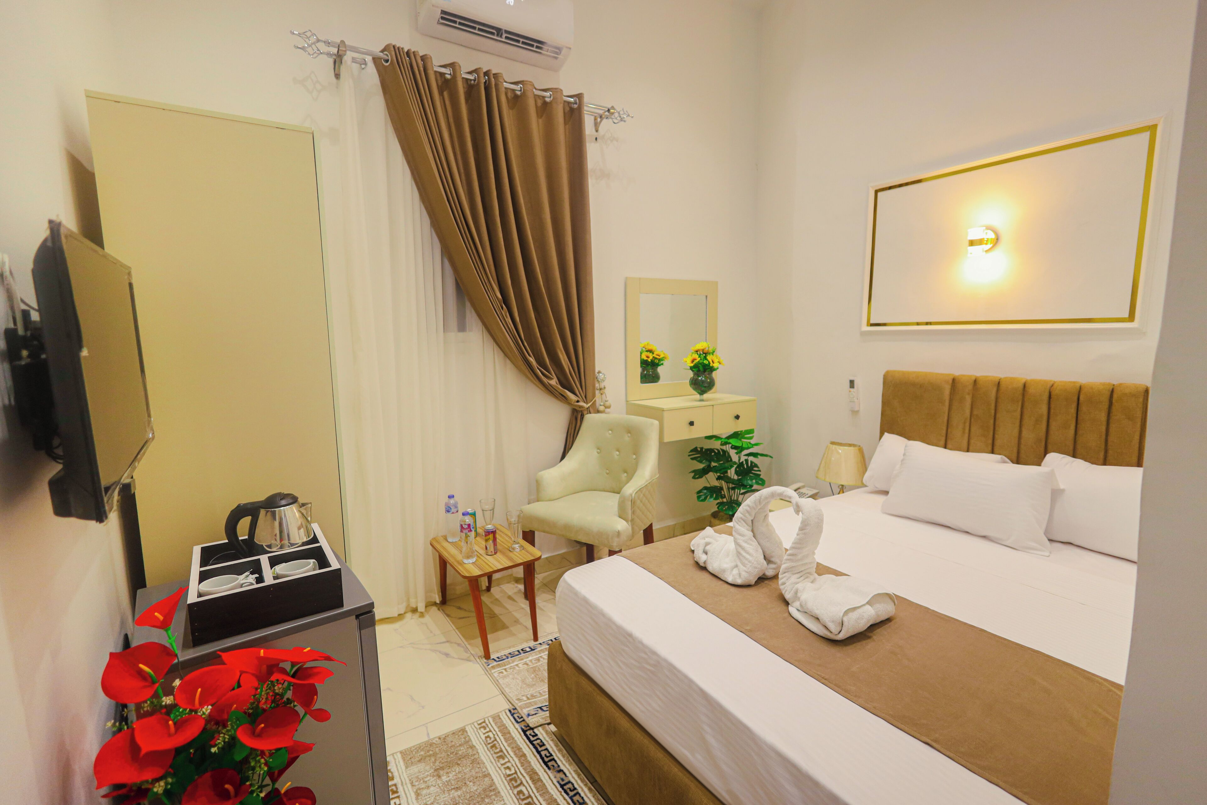 A hotel room with a bed, a chair, a desk with a kettle, a mirror, and a wall-mounted light fixture.