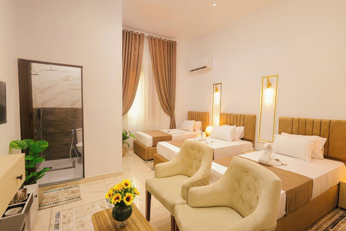 A hotel room with two beds, a sitting area with chairs, a small table with a vase of flowers, and a bathroom with a shower and toilet.