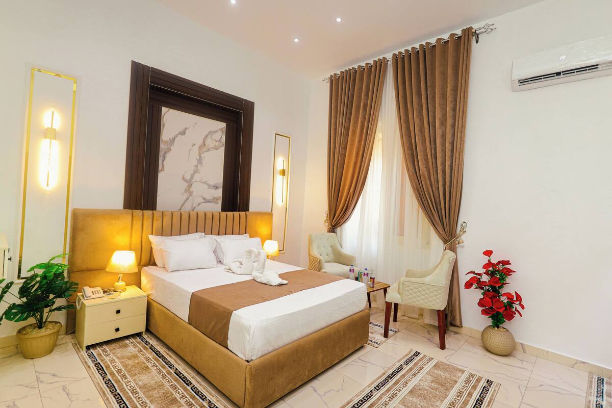 A hotel room with a bed, bedside lamps, a nightstand, a potted plant, a chair, and a vase with flowers.