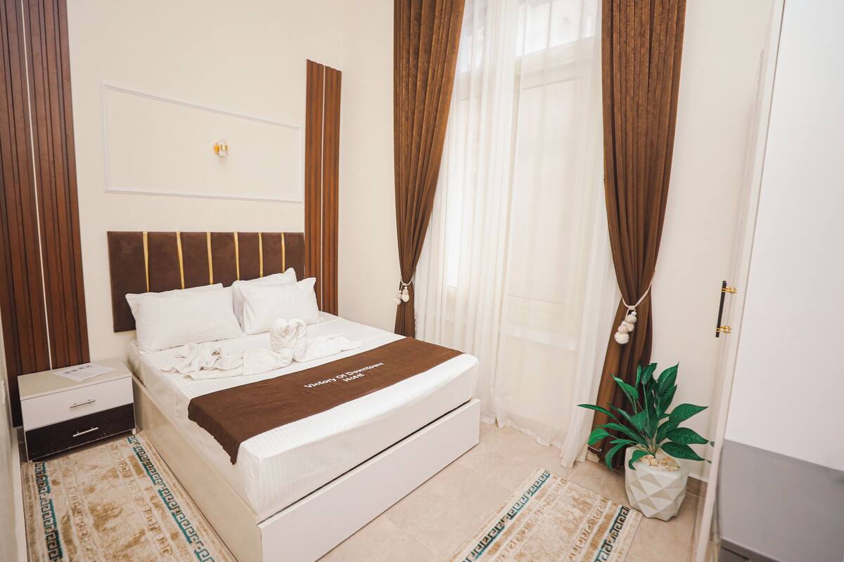 A neatly made bed with a brown blanket, a bedside table, a nightstand, a potted plant, and a window with curtains.