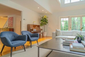 A modern living room with a piano, two blue armchairs, a white sofa, and a large window with a view of trees. - Southampton Estate w Heated Pool (Southampton)