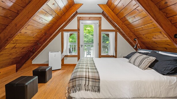 A cozy attic bedroom with a bed, two windows, a heater, and wooden beams.