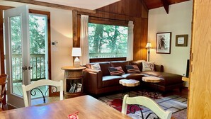 A cozy living room with a wooden ceiling, a brown leather sofa with American flag pillows, a wooden coffee table, and a dining table with chairs.