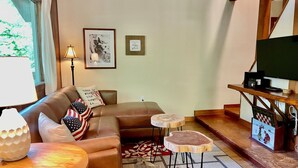 A cozy living room with a brown leather sofa, a small round table, a television on a wooden stand, and a framed picture on the wall.