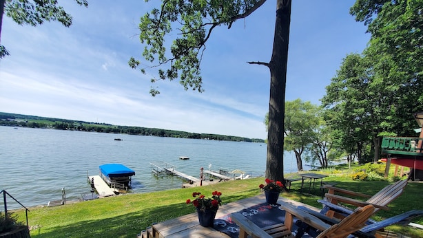 A wooden deck with a table and chairs overlooking a lake, a dock with a boat, and a building with a green roof. - Lakefront cottage on Silver lake near Letchworth Park (Castile)
