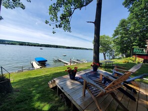 A wooden deck with a table and chairs overlooking a lake, a dock with a boat, and a building with a green roof. - Lakefront cottage on Silver lake near Letchworth Park (Castile)