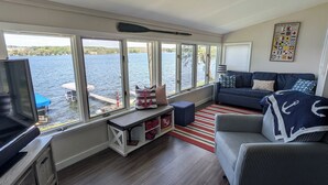 A living room with a large window overlooking a body of water, a television, a sofa, and a nautical-themed wall decoration. - Lakefront cottage on Silver lake near Letchworth Park (Castile)