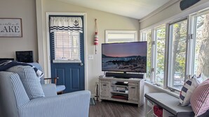 A living room with a blue sofa, a television, a window with a view of trees, and a striped rug. - Lakefront cottage on Silver lake near Letchworth Park (Castile)