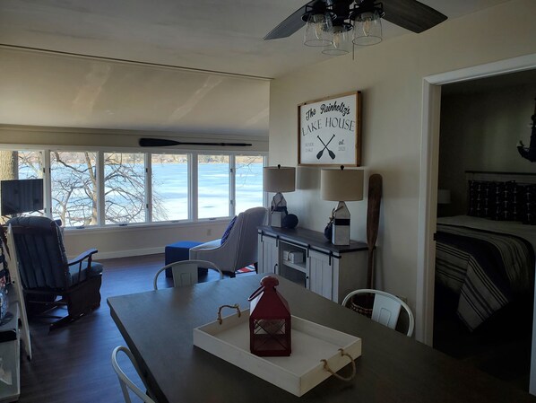A room with a dining table, chairs, a TV, and a large window with a view of a lake. - Lakefront cottage on Silver lake near Letchworth Park (Castile)
