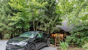 A Tesla parked in front of a rustic cabin surrounded by lush greenery. - Bear Pause Cabin 3 BR/3BA Amazing Lake View EV Charger (Seven Devils)