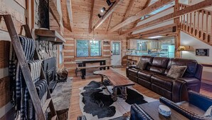 A rustic cabin interior with exposed wooden beams, a leather sofa, a dining table, and a stone fireplace. - Bear Pause Cabin 3 BR/3BA Amazing Lake View EV Charger (Seven Devils)