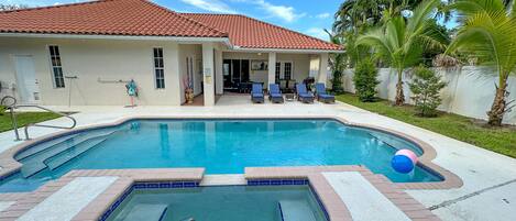 A swimming pool with a tiled deck, a covered patio area with lounge chairs, and palm trees in the background.