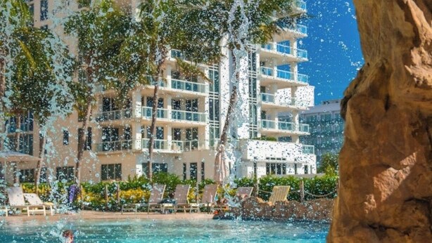 A hotel pool with clear water, surrounded by palm trees and a large building in the background.