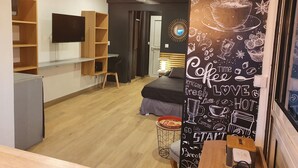 Une chambre moderne équipée d’un lit, d’un bureau et d’une télévision. Un des murs est décoré sur le thème du café et le plafond est doté d’un ventilateur.