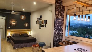 Une chambre à coucher moderne comprenant un lit, des tables de chevet, un petit tabouret, un mur orné de tableaux encadrés, un panneau décoratif mural et une fenêtre donnant sur un paysage.