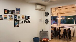 Une pièce comprenant un lit, un coin repas avec une table et des chaises, un fauteuil gris, une petite table ronde avec un plateau, et un mur orné de tableaux et d’une horloge.