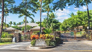 Une entrée fermée par un portail, agrémentée d’une enseigne accueillant les visiteurs aux Palm Villas, le tout étant entouré d’une végétation tropicale luxuriante et de palmiers.