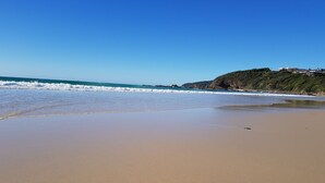 A sandy beach with gentle waves, a clear blue sky, and a rocky cliff with buildings in the background.