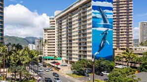 Un grand hôtel avec de nombreuses étages, orné d’une immense fresque représentant une baleine sur l’un de ses murs, une rue animée où circulent des voitures, et des palmiers.
