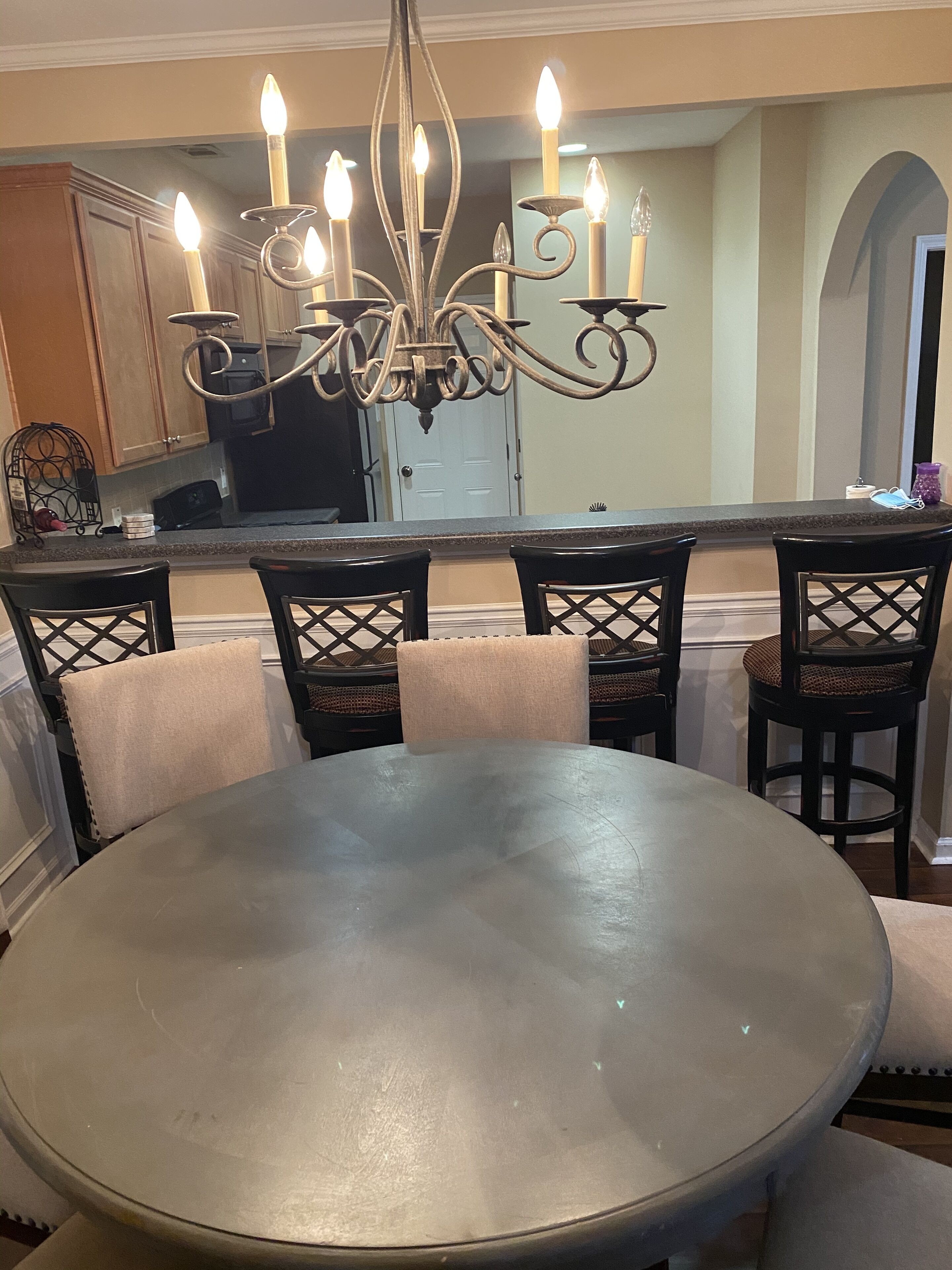 A dining area with a round table, six chairs, a chandelier with multiple candles, and a kitchen in the background.
