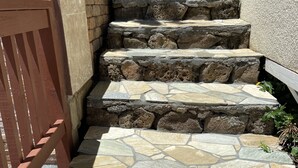 Eine Treppe aus Stein führt zu einer Tür, links ein Holzzaun und rechts eine Steinmauer.