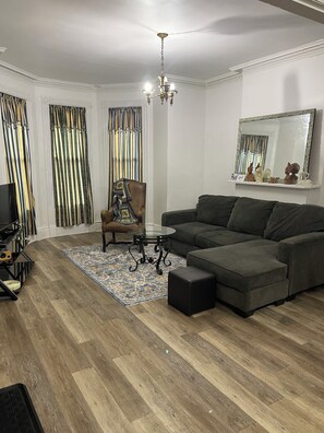Una sala de estar con un sofá modular, una mesa de centro de vidrio, un tapete estampado, una silla y un televisor.