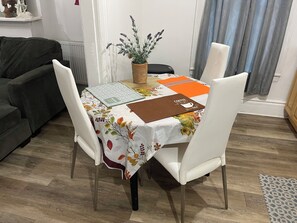 Una mesa de comedor con mantel floral, sillas blancas, una planta en maceta y una libreta marrón.