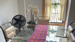 Una sala de conferencias con una mesa de vidrio, sillas de cuero, un ventilador, un florero con flores blancas y una ventana con cortinas.