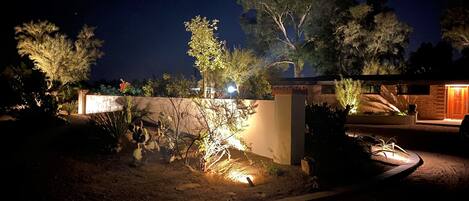 A nighttime view of a residential property with a well-lit entrance, a paved driveway, and a variety of plants and trees.
